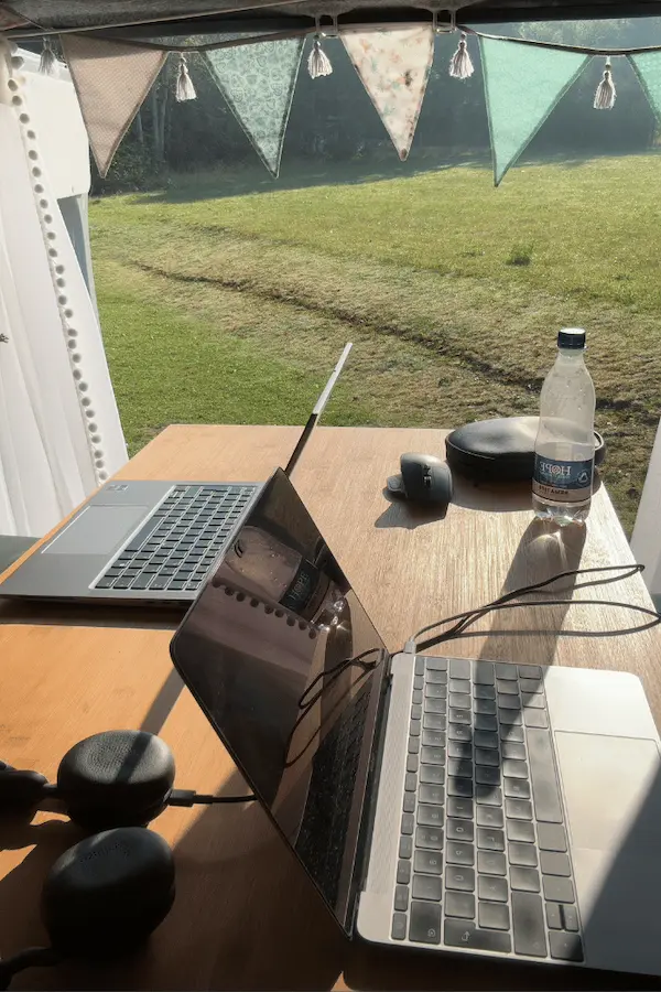 Camper Office - zweit aufgeklappte Laptops stehen auf einem Tisch im hinteren Bereich eines Campervans. Die Türen sind offen und man sieht auf eine Wiese