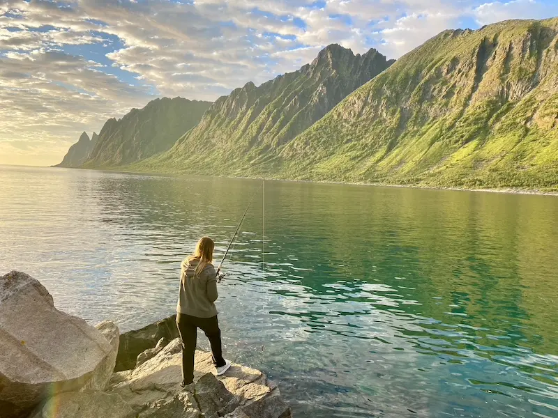 Frau steht auf einem Stein mit einer Angel in der Hand. Im Hintergrund sind die Berge von Senja zu sehen, die Sonne geht gerade unter. Vanlife-Tipp: Hobby suchen